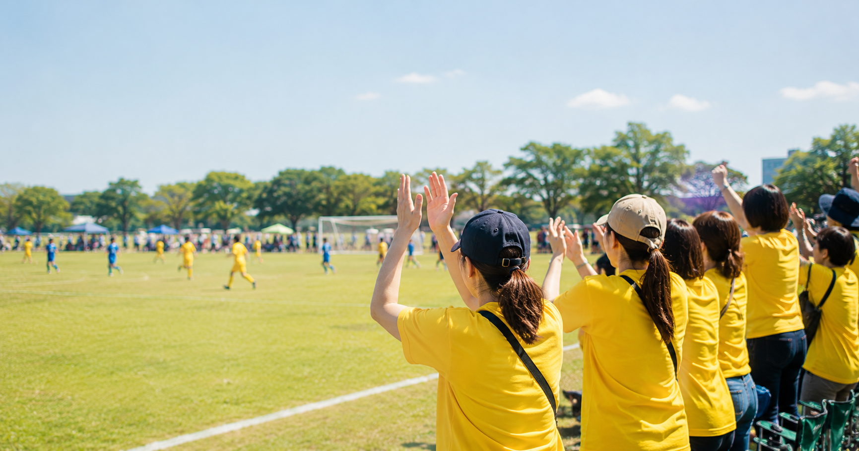 少年サッカーの試合会場でお揃いの黄色い応援Tシャツを着て声援を送る保護者たち